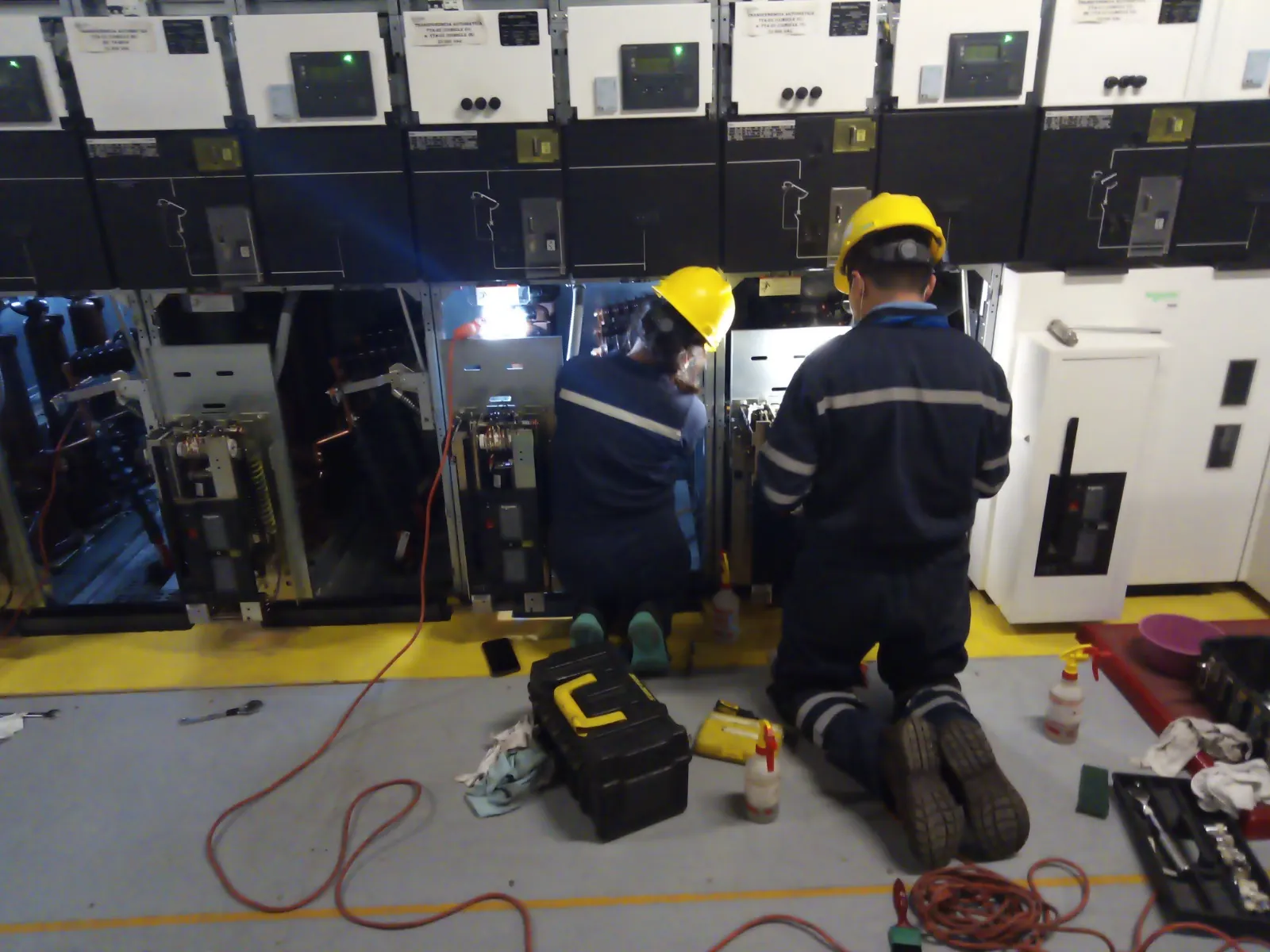 Dos técnicos con casco amarillo y overol trabajando de rodillas frente a celdas de media tensión